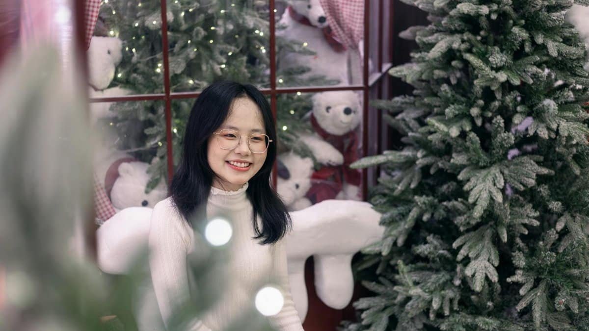 Woman smiling surrounded by Christmas decorations and pine trees indoors. Festive and joyful holiday setting.
