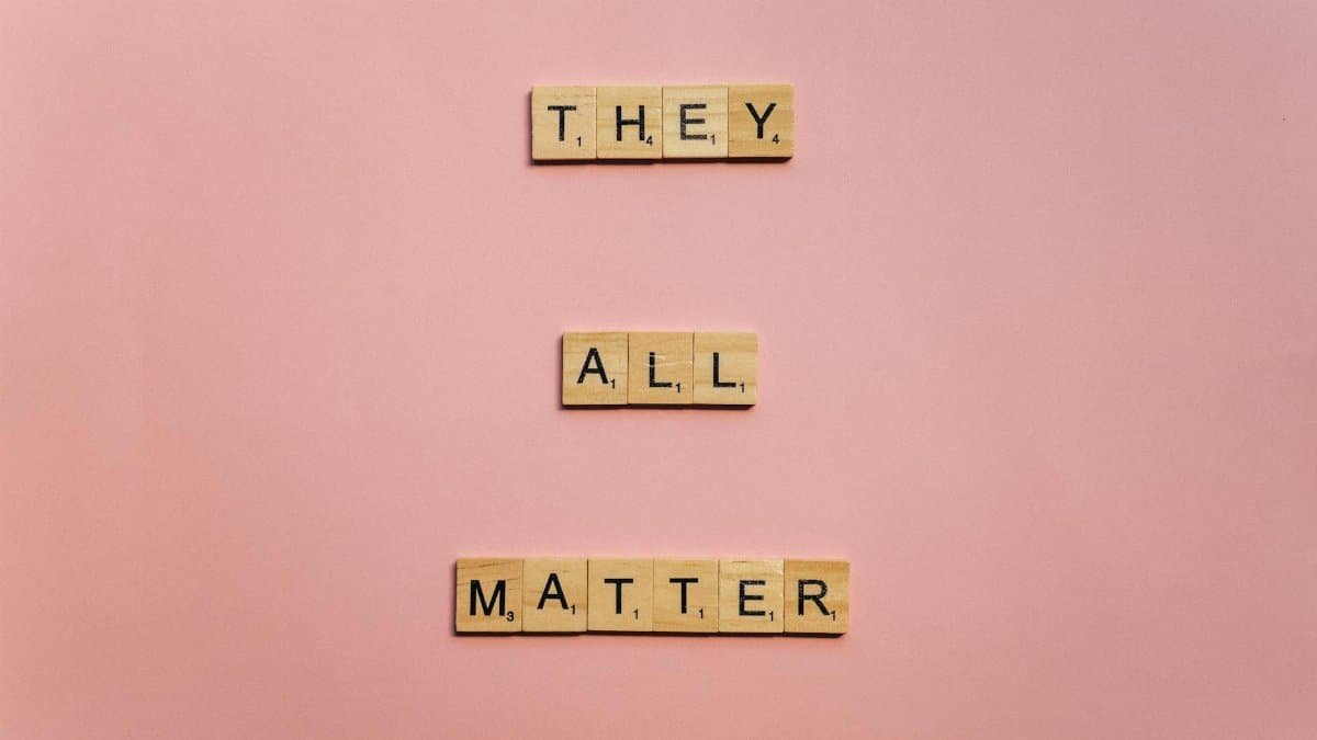 Wooden Scrabble tiles spell 'They All Matter' on a pink background, promoting inclusivity.