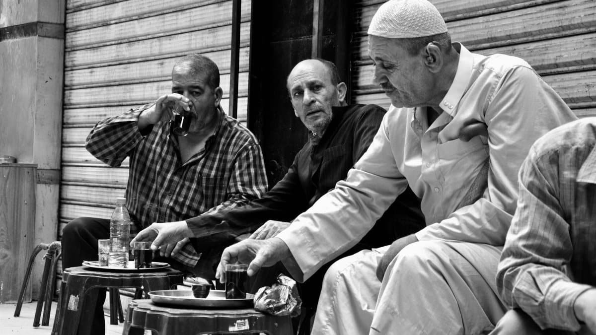 A candid black and white photo of three middle-aged men enjoying tea in an outdoor market setting.