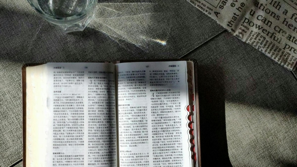 Sunlit open book and glass on a table with newspaper, creating a serene still life scene.