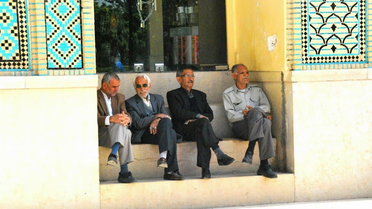 A group of four middle-aged and senior men sitting outside in Shiraz, showcasing traditional Iranian architecture.
