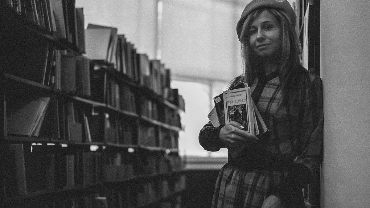 Black and white portrait of a woman in a library holding books, wearing a beret.