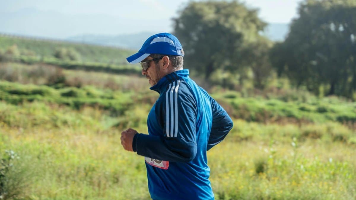 Middle-aged man in blue sportswear jogging on a sunny outdoor trail surrounded by greenery.