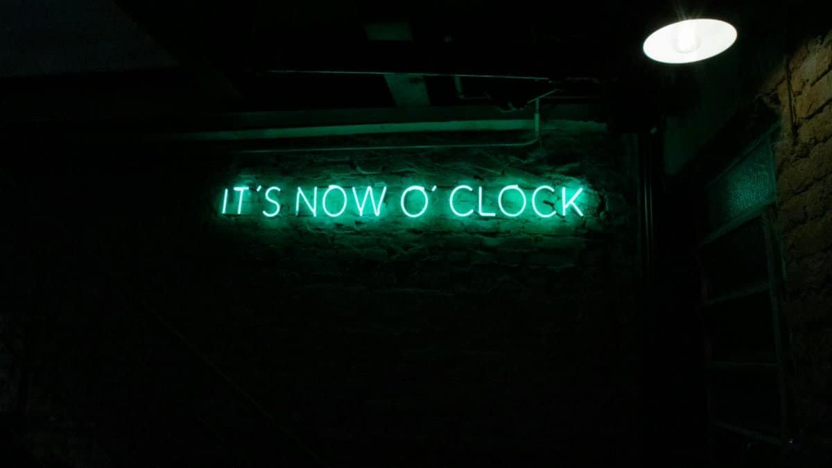 Green neon sign in a dimly lit room showcasing 'It's Now O' Clock'.