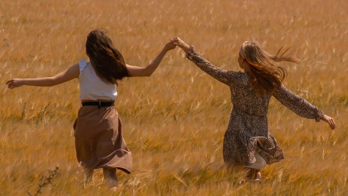 Two women run joyfully holding hands through a sunlit wheat field.