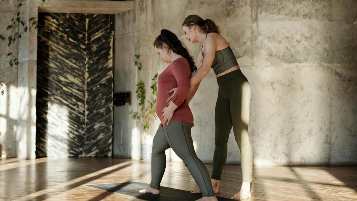 A woman supports another woman with Down syndrome during a yoga session indoors.