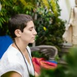 A person engaged in serene meditation outdoors near a Buddha statue amidst lush greenery.