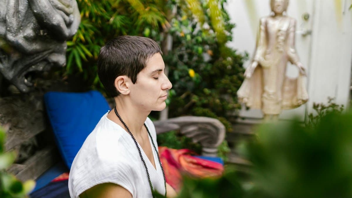 A person engaged in serene meditation outdoors near a Buddha statue amidst lush greenery.
