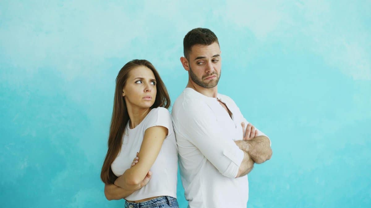 Young couple stands back to back with crossed arms against blue background.