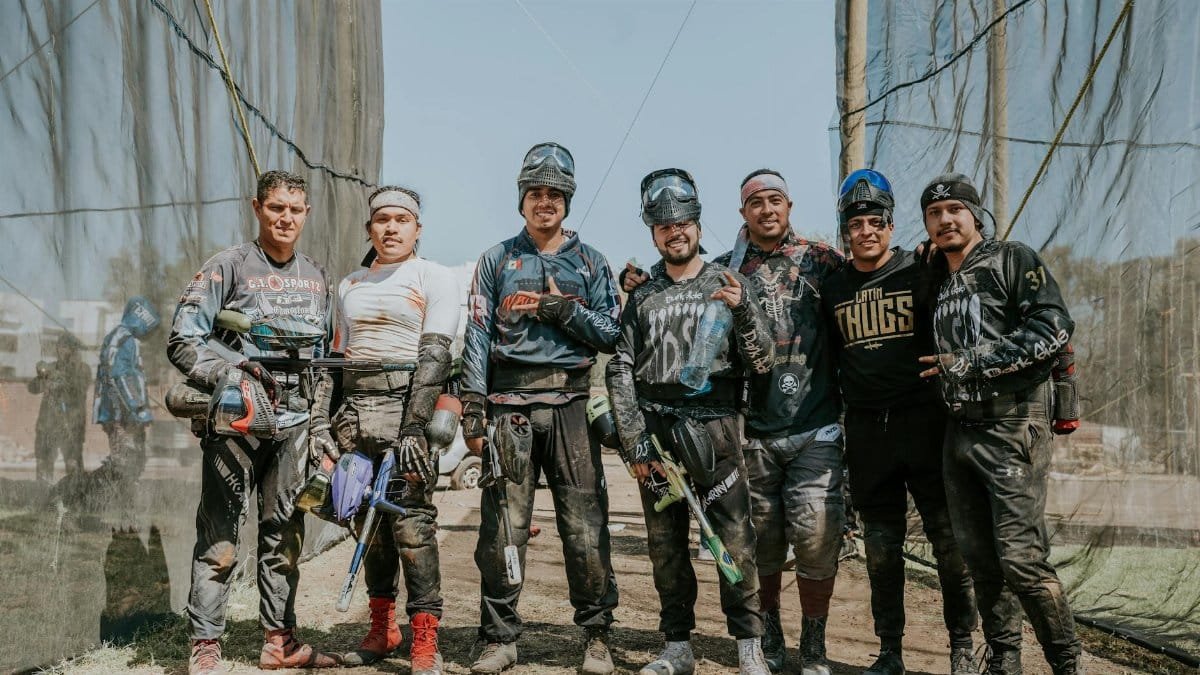 A team of men in paintball gear posing outdoors with smiles and paint markers.