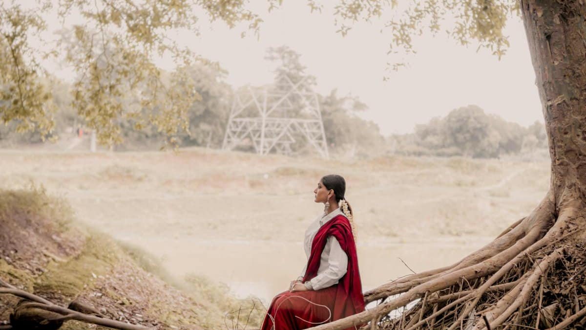 A serene portrait of a woman in a red saree sitting by tree roots along a riverside in Varanasi, India.