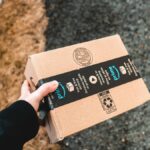 A person holding a cardboard Amazon Prime package on a snowy urban sidewalk.