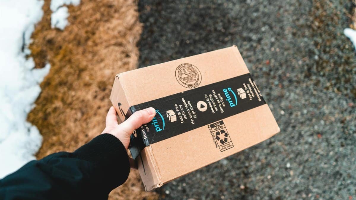 A person holding a cardboard Amazon Prime package on a snowy urban sidewalk.