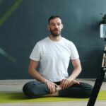 A man in white T-shirt filming an online yoga session indoors using a smartphone on a tripod.