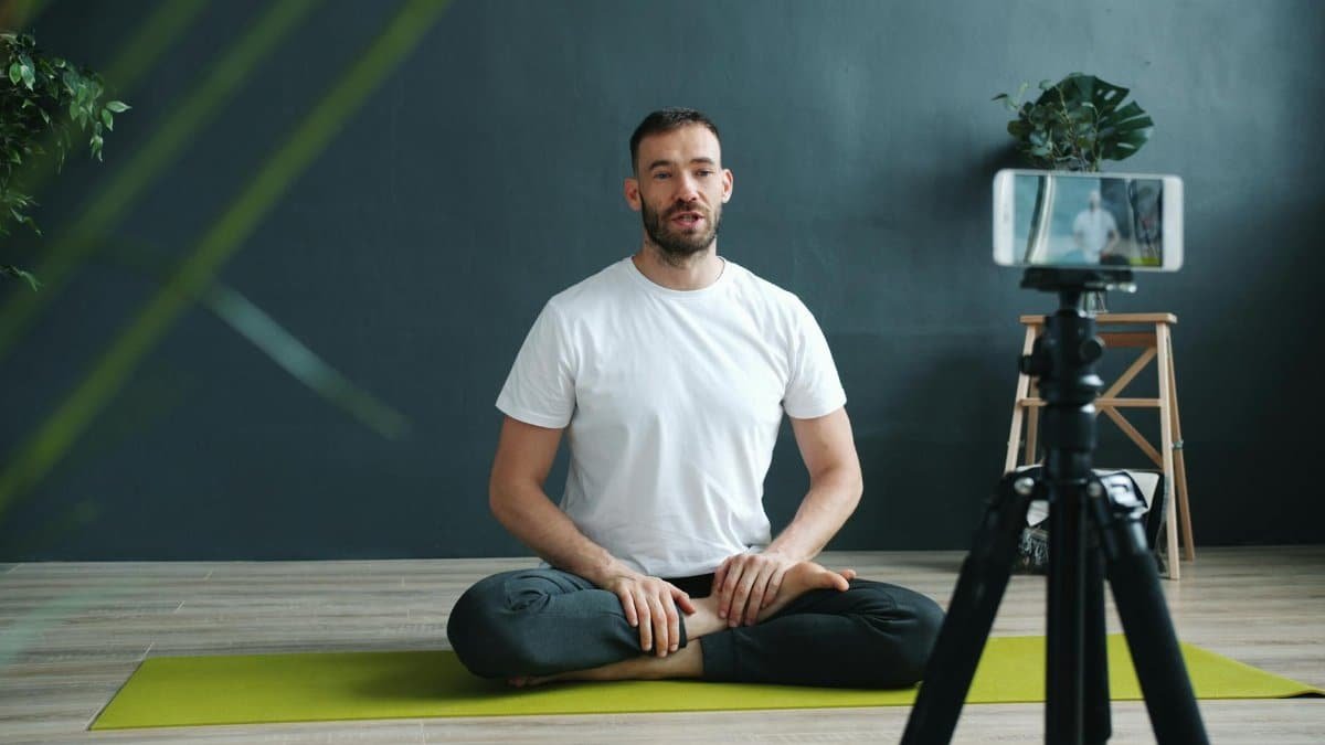 A man in white T-shirt filming an online yoga session indoors using a smartphone on a tripod.