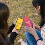 Two women using dating apps Bumble and Tinder on smartphones in a park setting.