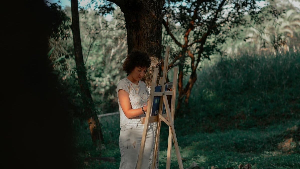 A woman artist painting on a canvas outdoors in a serene forest environment.