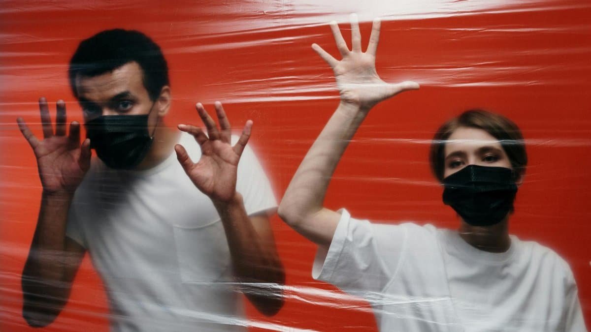 Two people wearing masks stand behind a plastic film against a red background.