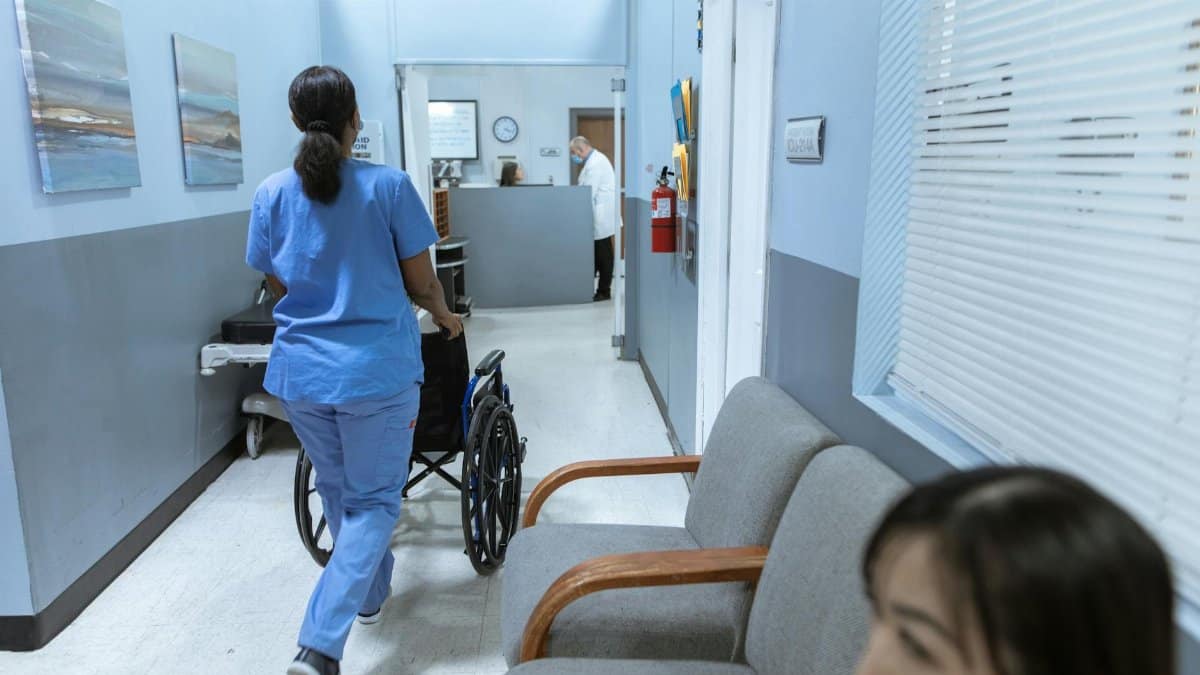 A nurse in scrubs pushes a wheelchair through a well-lit hospital corridor, attending to patient care.