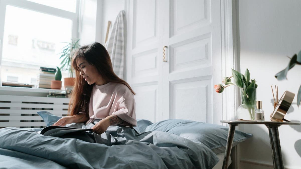 A serene morning scene with an Asian woman reading in a sunlit bedroom.