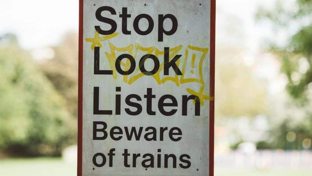 Rectangular signboard with Beware Of Trains title on post near city park in daytime