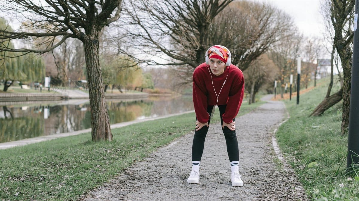 A woman in a head scarf and headphones rests on a tree-lined riverside path during autumn.
