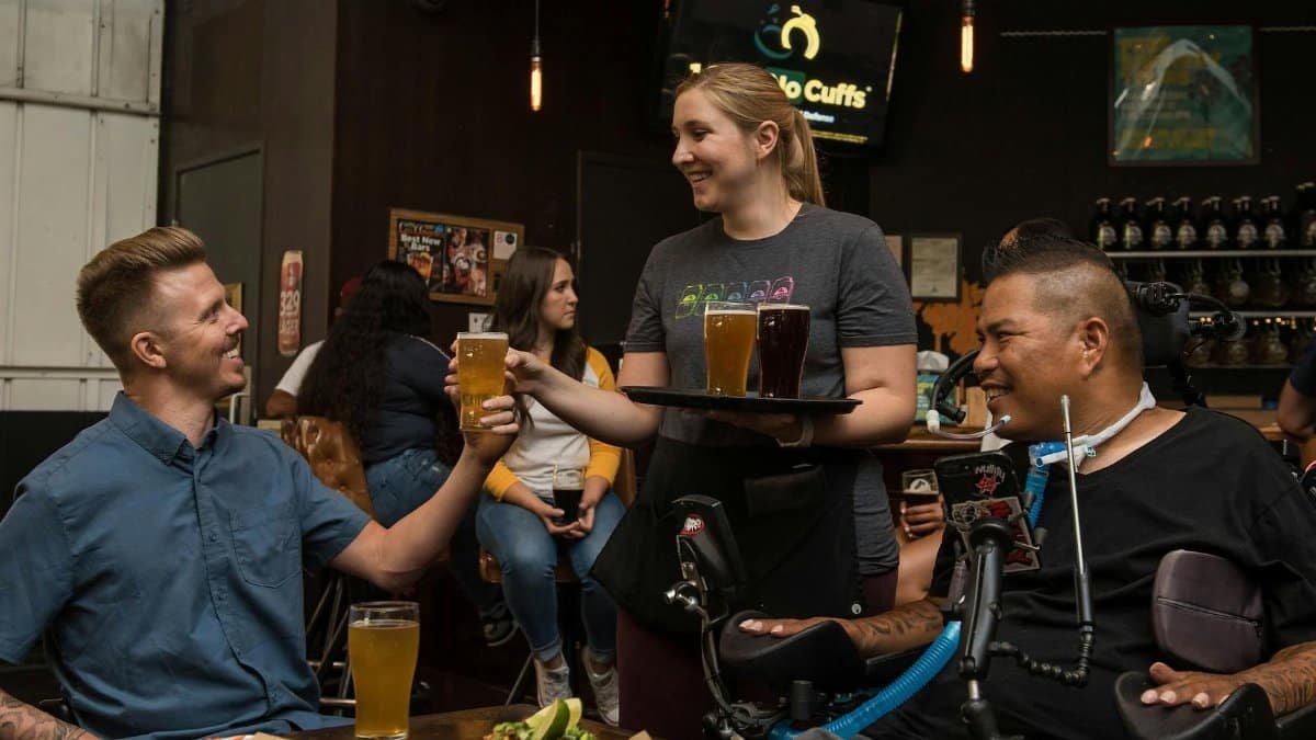 A waitress serves drinks to friends, including one in a wheelchair, in a lively bar setting.