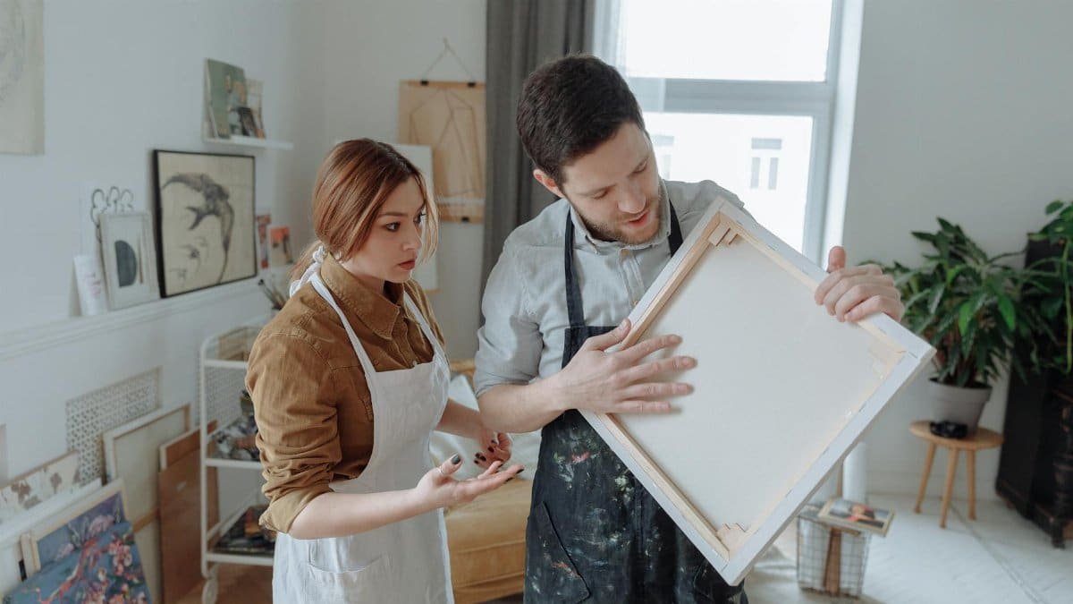 Two artists in aprons discuss art techniques with a blank canvas in a creative studio.