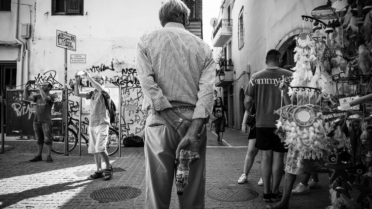 Monochrome street scene capturing an elderly man with dreamcatchers and graffiti in the background.