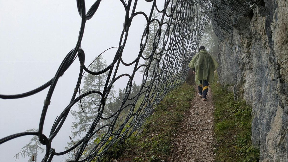 A solitary hiker walks along a misty mountain trail secured with protective netting.