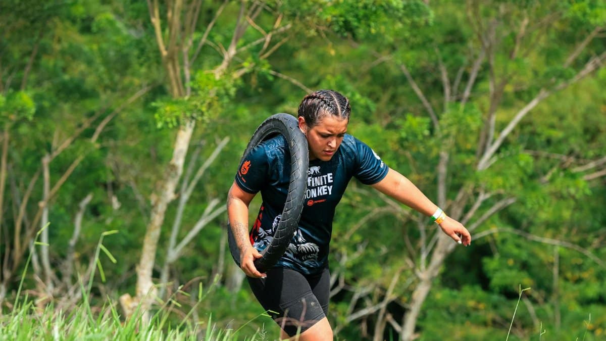 A woman carries a tire outdoors during a fitness challenge surrounded by greenery.