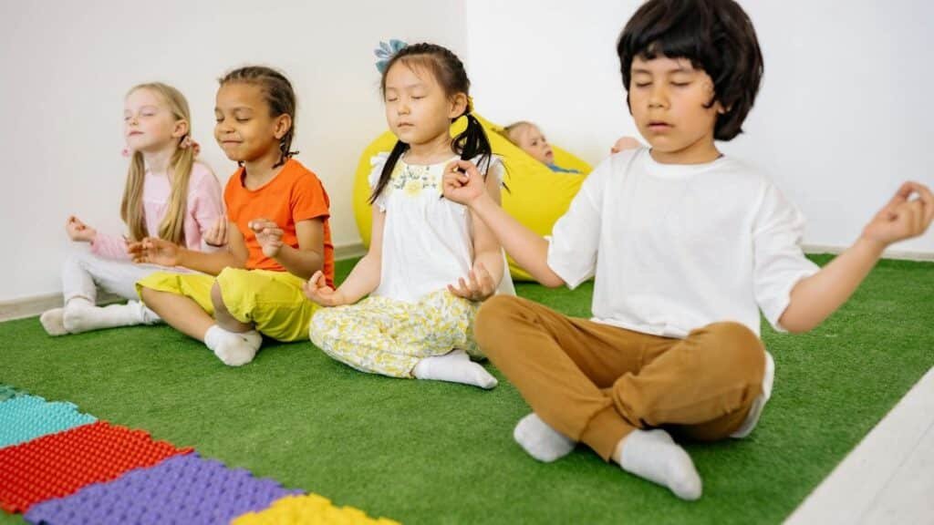 Group of diverse young children meditating in yoga poses on a green mat indoors.