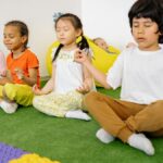 Group of diverse young children meditating in yoga poses on a green mat indoors.