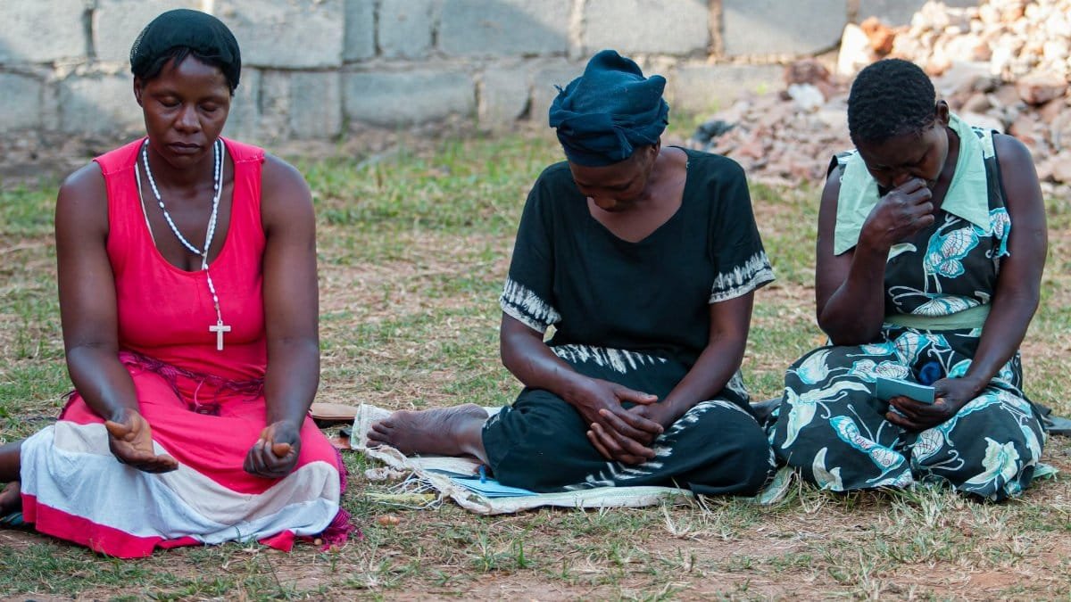 Three women sitting and praying outdoors, expressing peace and contemplation.