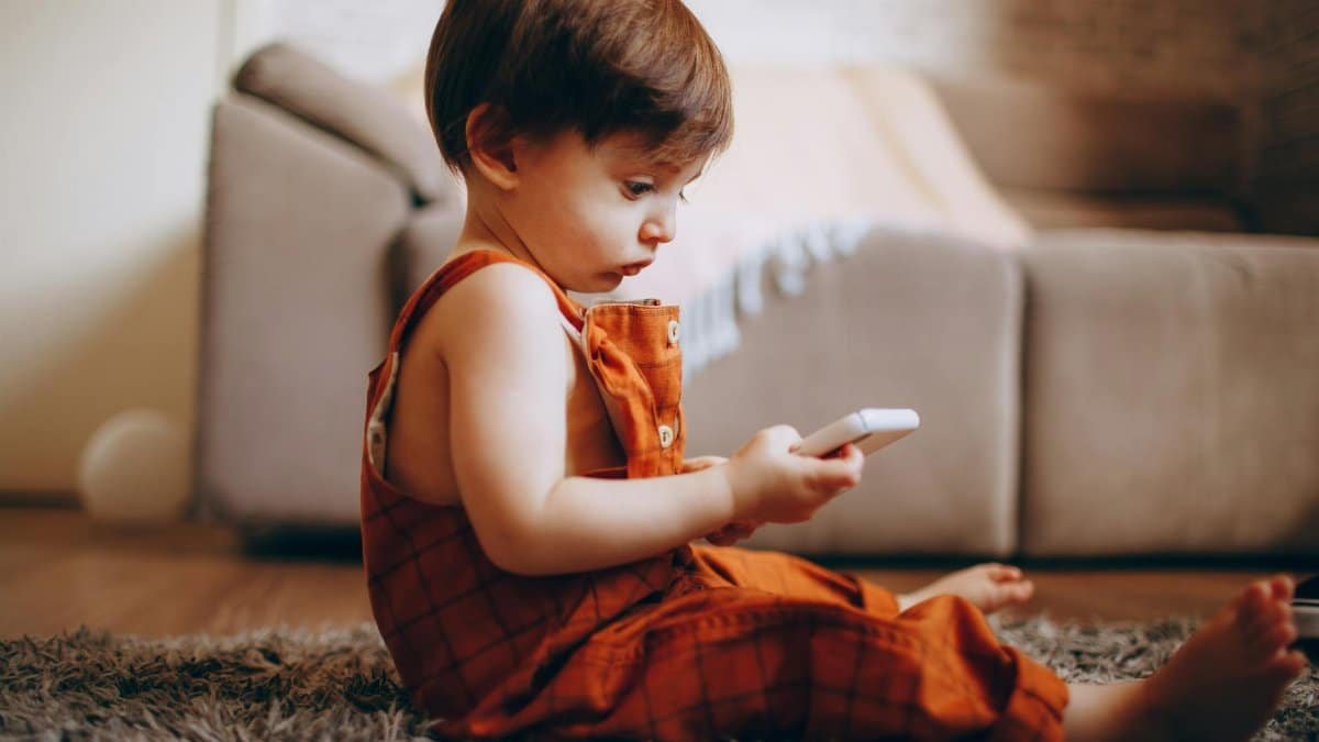 A cute toddler in orange overalls sits on the carpet, focused on a smartphone, indoors.