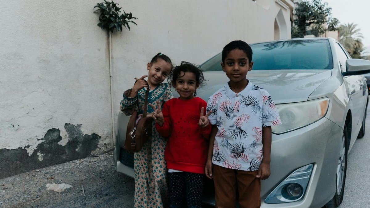 Three children posing happily by a car in Nizwa, Oman showcasing local culture and architecture.