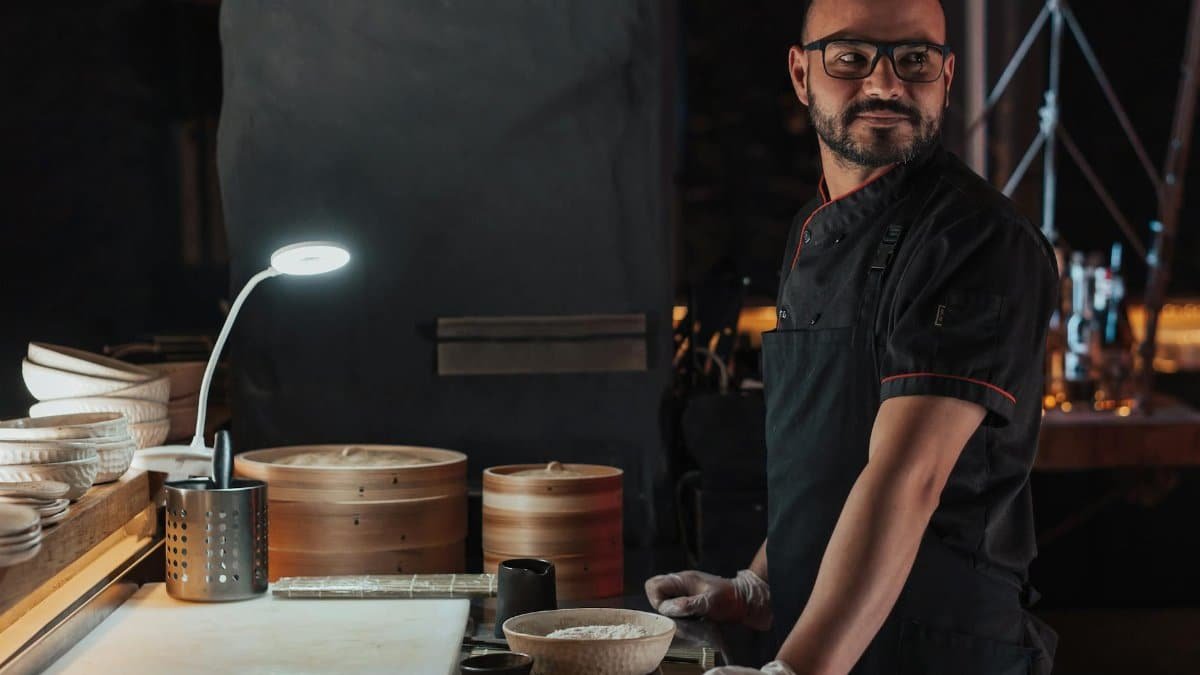 A professional chef stands confidently in a dimly lit kitchen with bamboo steamers and culinary tools.