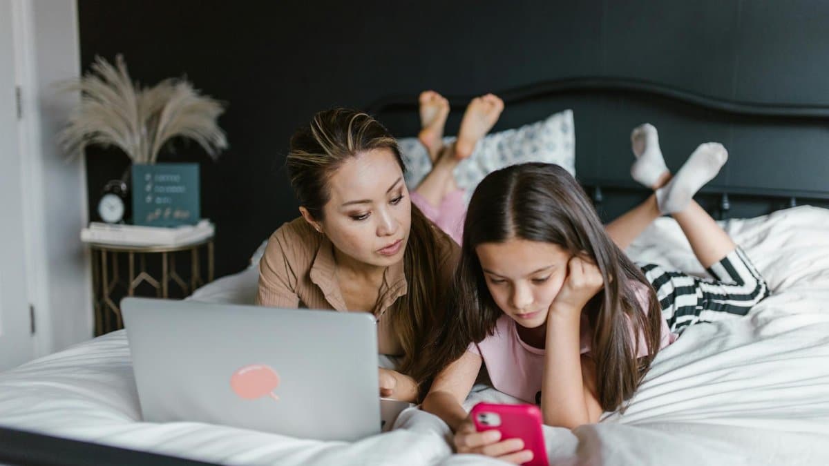 A mother and daughter are focused on electronic devices while lying on a bed, reflecting modern family life.
