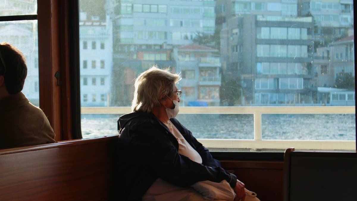 A senior woman wearing a mask gazes out the ferry window, reflecting by the sea.