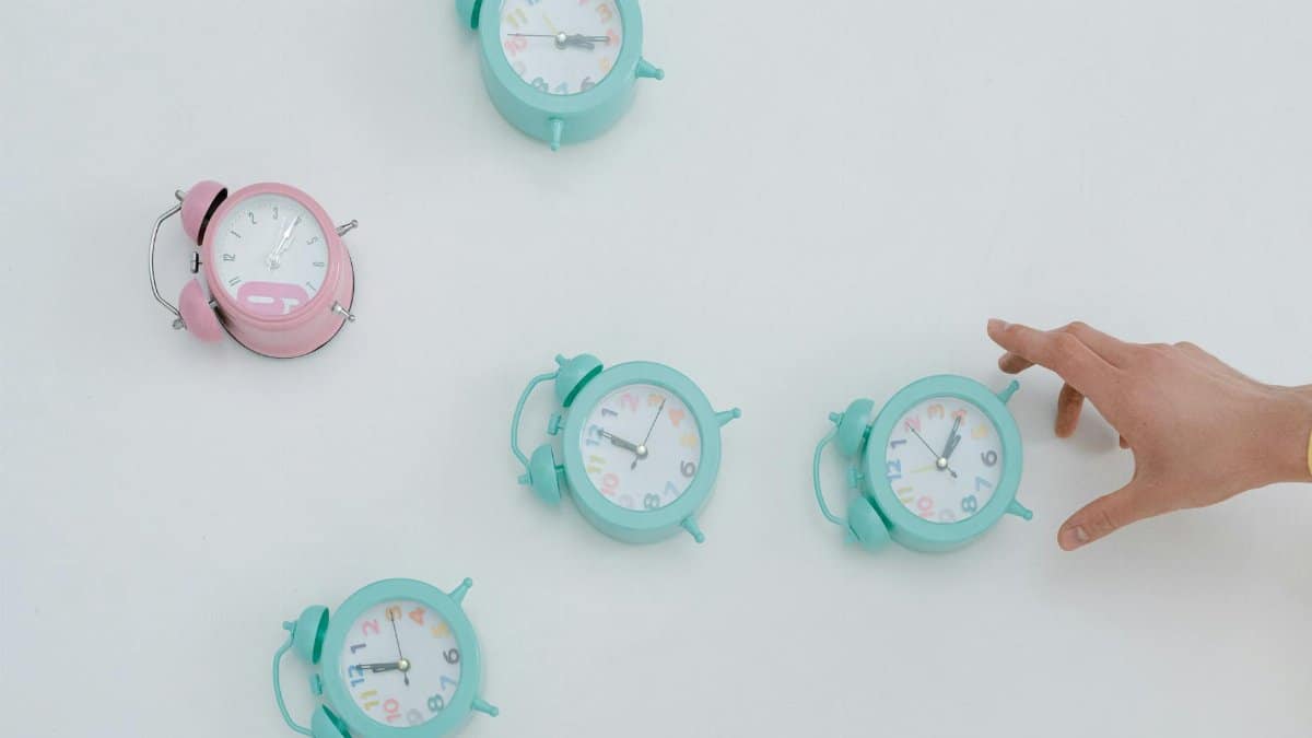 A hand reaching for pastel alarm clocks on a plain white background. Minimalist style.