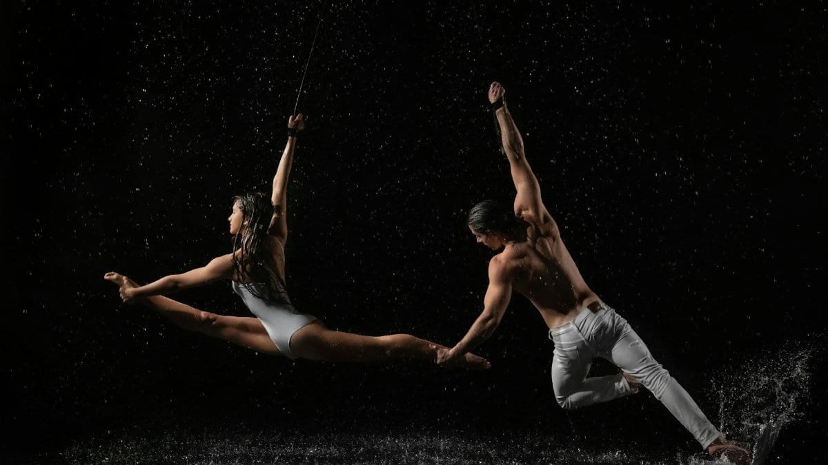 Male and female dancers perform a dynamic leap with water splashes, evoking movement and drama.