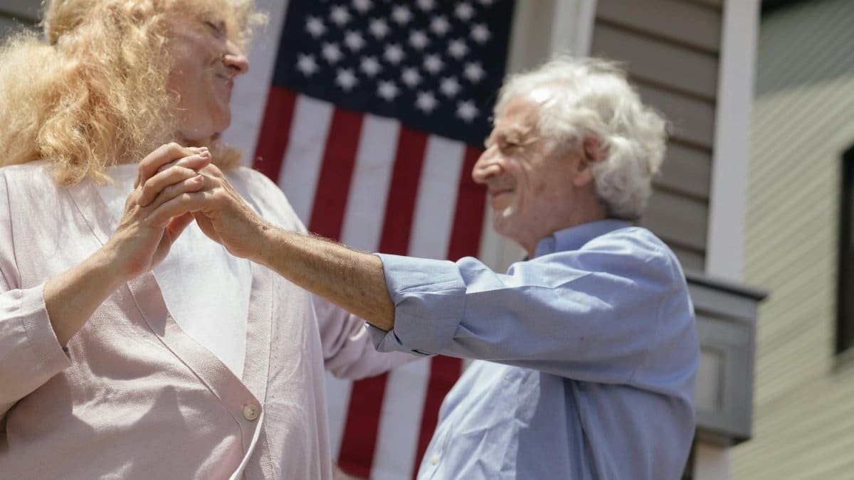 Smiling senior couple dancing with an American flag background, celebrating Independence Day.
