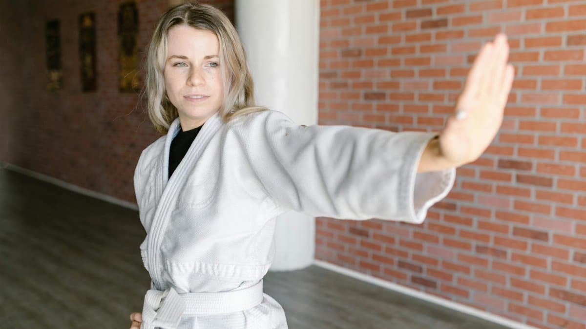 A woman in a martial arts uniform practices a defensive stance indoors against a brick wall.