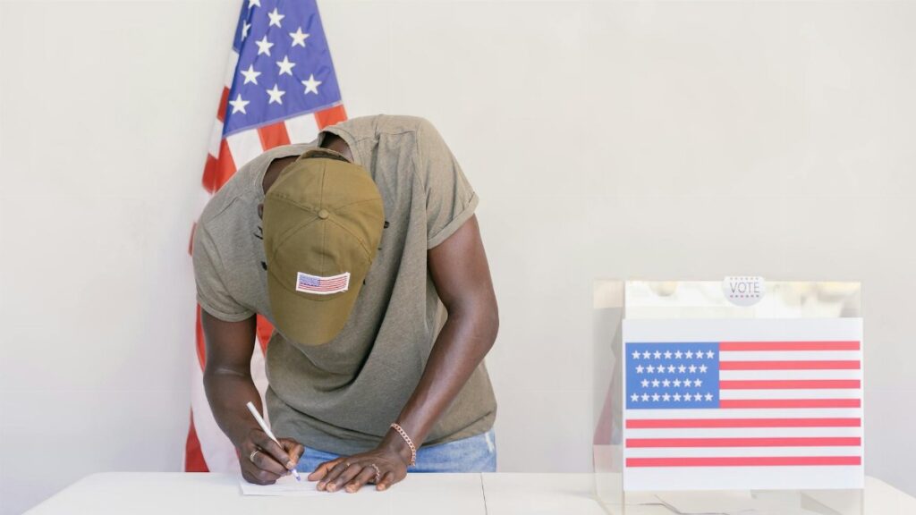 Man casting vote at polling station with American flag in background.