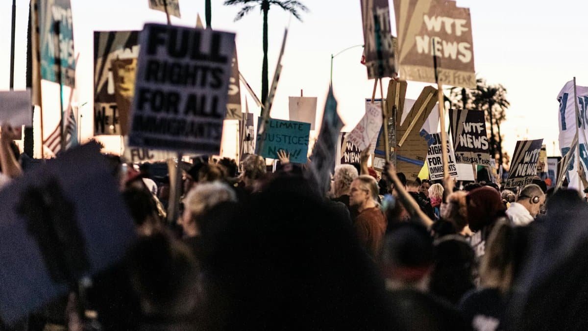 A diverse crowd gathers outdoors holding protest banners and signs at dusk.