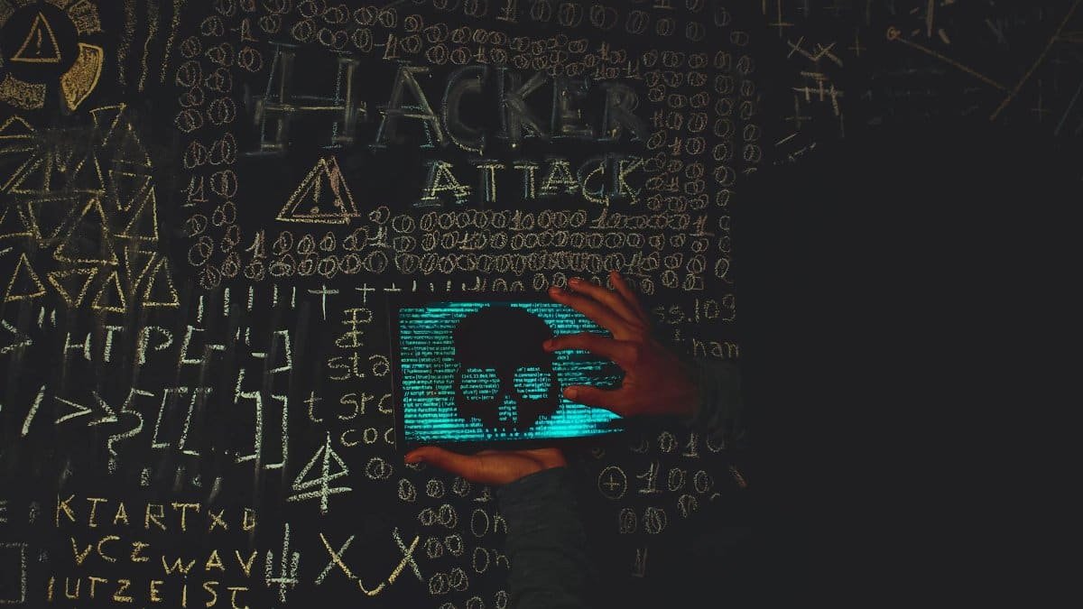 A hacker in a black hoodie using a tablet displaying a skull, surrounded by chalk symbols and 'Hacker Attack' text.