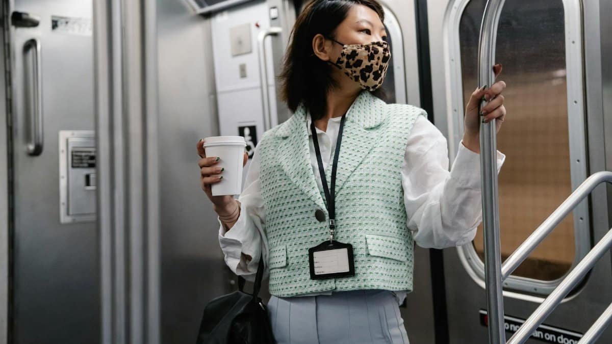 Asian woman wearing a facemask holding coffee on a subway train, depicting the new normal of commuting.