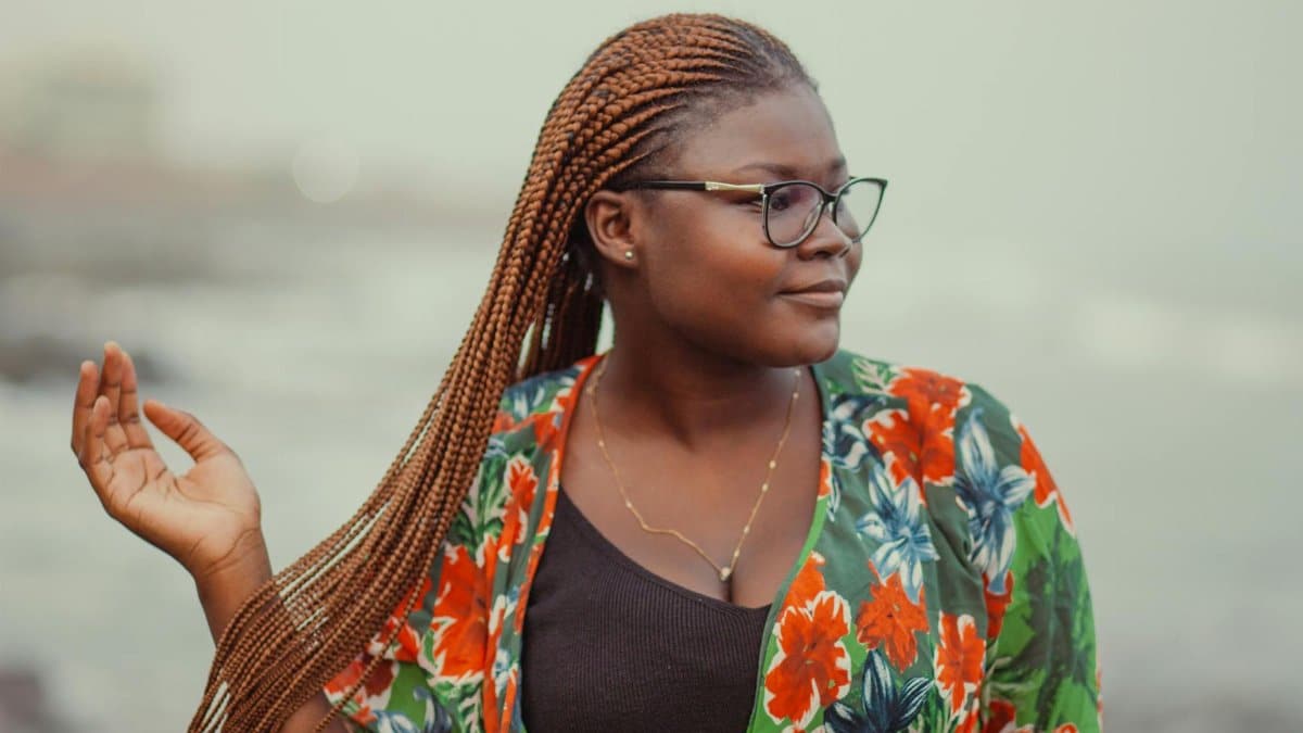 Self assured young black female tourist in fashionable outfit and eyeglasses touching long Afro braids and looking away while relaxing on seashore
