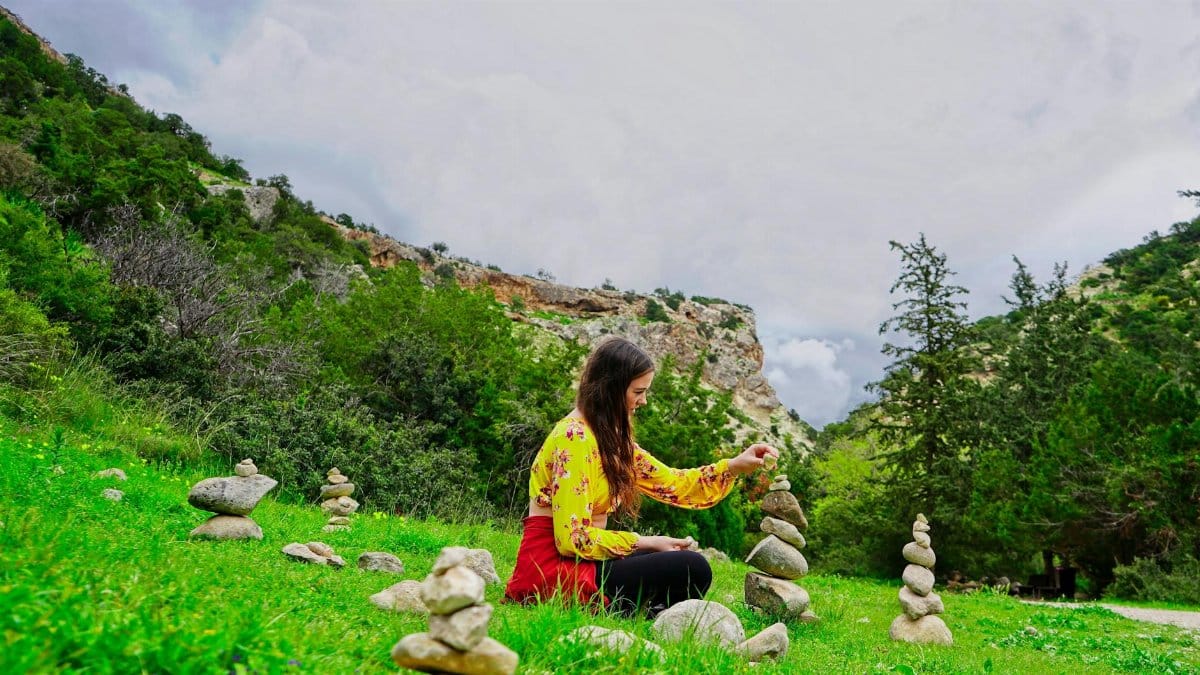 Side view of tranquil female with long hair in colorful clothes sitting on ground and putting rocks on top of each other while meditate in nature in daylight