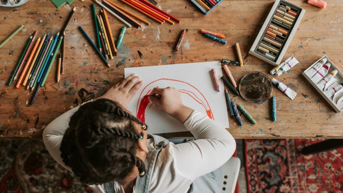 Child engaged in creative drawing with colored pencils, crayons, and paints at a wooden table.